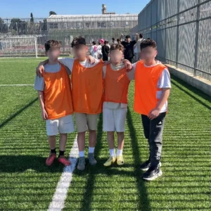 ארבעה ילדים באפודי אימון כתומים עומדים עם ידיים על הכתפיים על מגרש כדורגל מדשא.