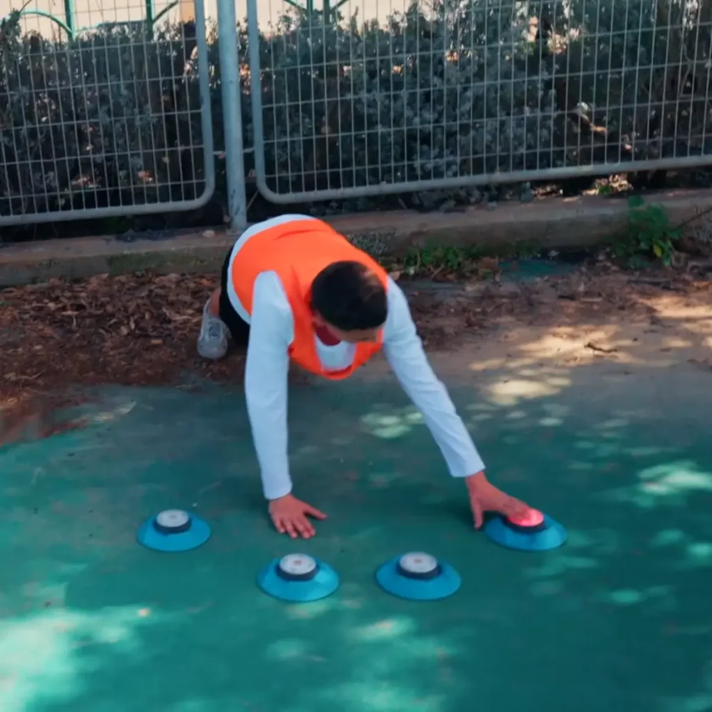 מתאמן באפוד כתום מבצע תרגיל פלאנק במגרש פתוח תוך כדי לחיצה על אורות אימון כחולים על הקרקע.